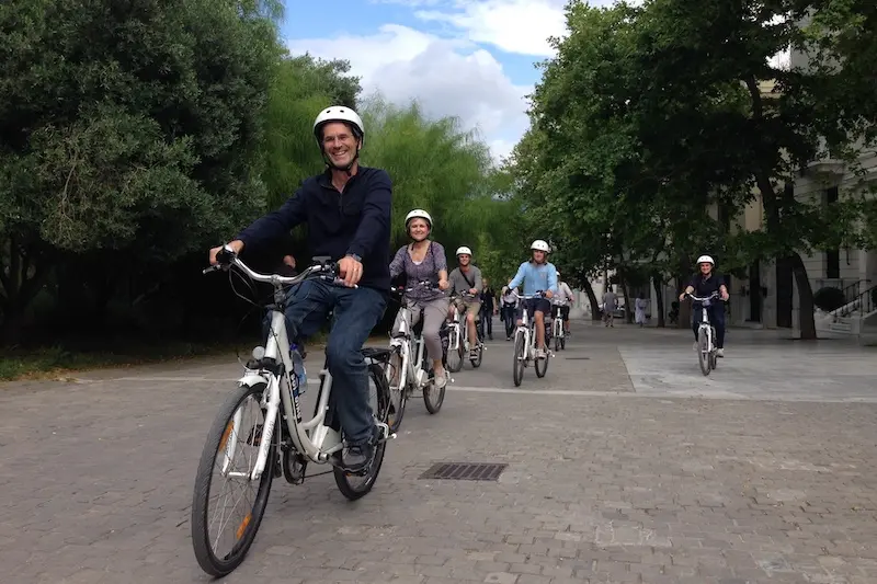 A family of adults riding electric bikes past the stone arches of the Herodion Theatre under the Acropolis in Athens.