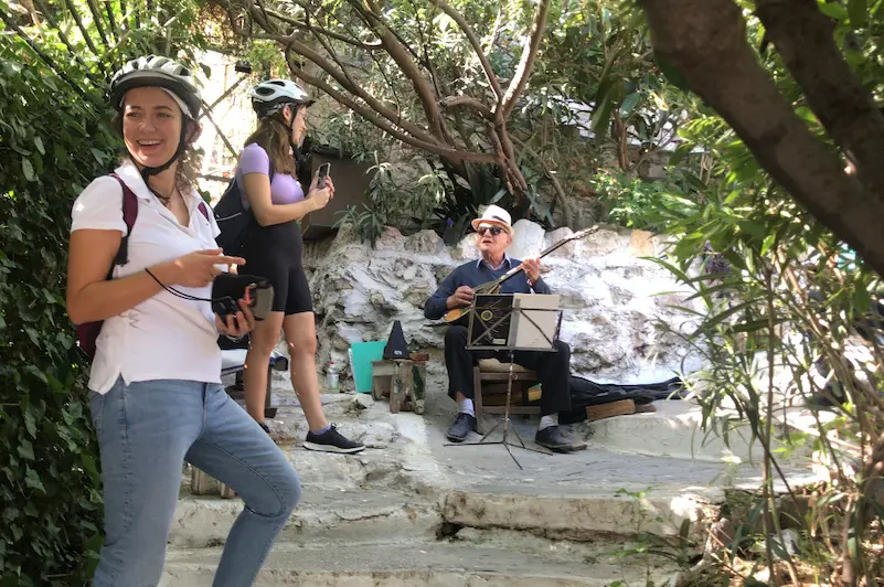 Female e-bike riders stopping to listen to a traditional Greek bouzouki player in a narrow Plaka alleyway, Athens.