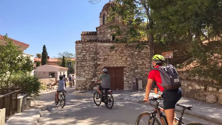 Riders cycling past a historic church near the north slope of the Acropolis in Athens